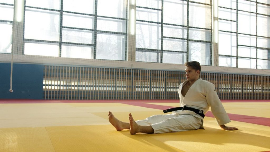 A martial artist in a dojo practices meditation on a sunlit mat.